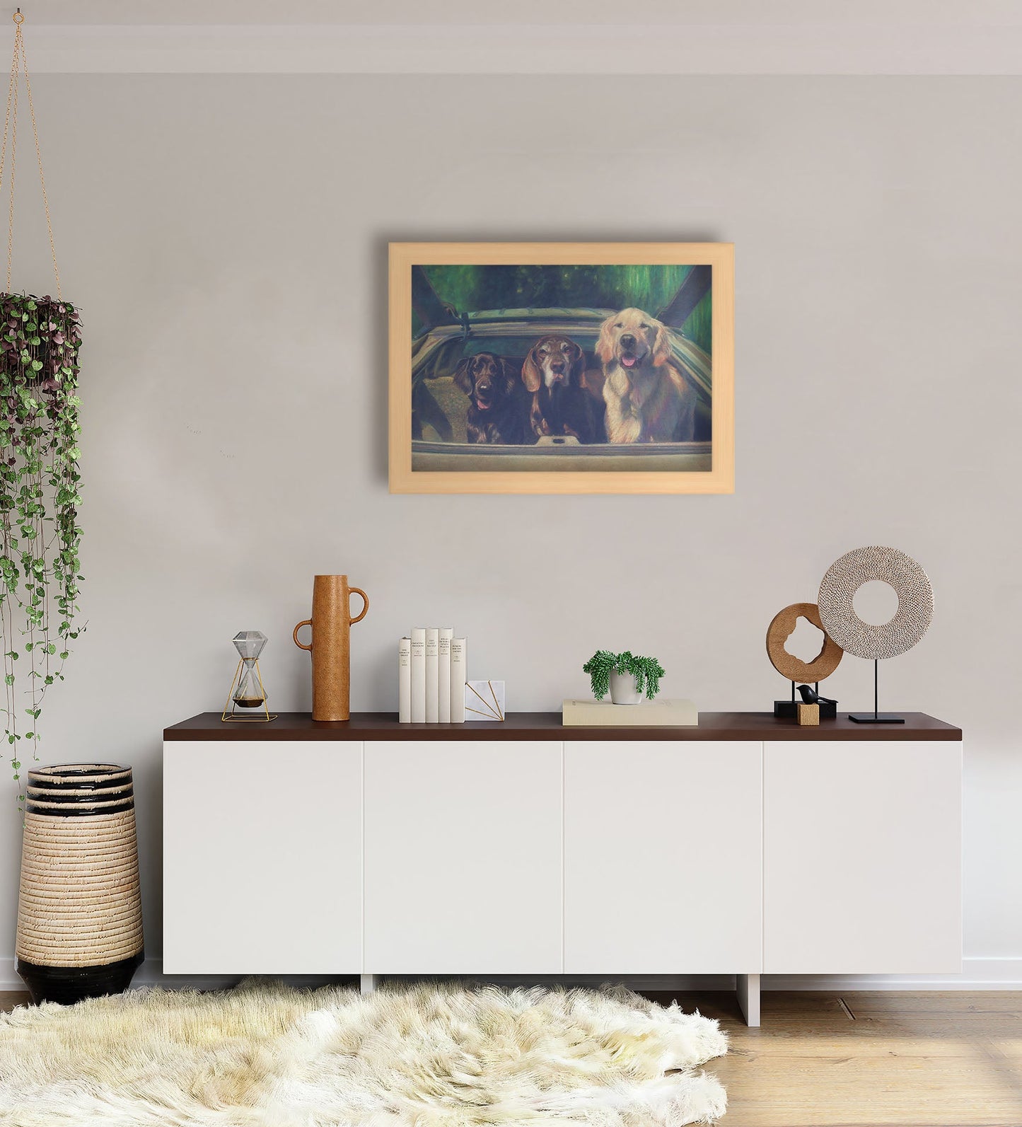 A white-walled room with a long, white cabinet decorated with sculptures, plants, and a woven basket. A framed painting of three dogs riding in a car hangs on the wall.