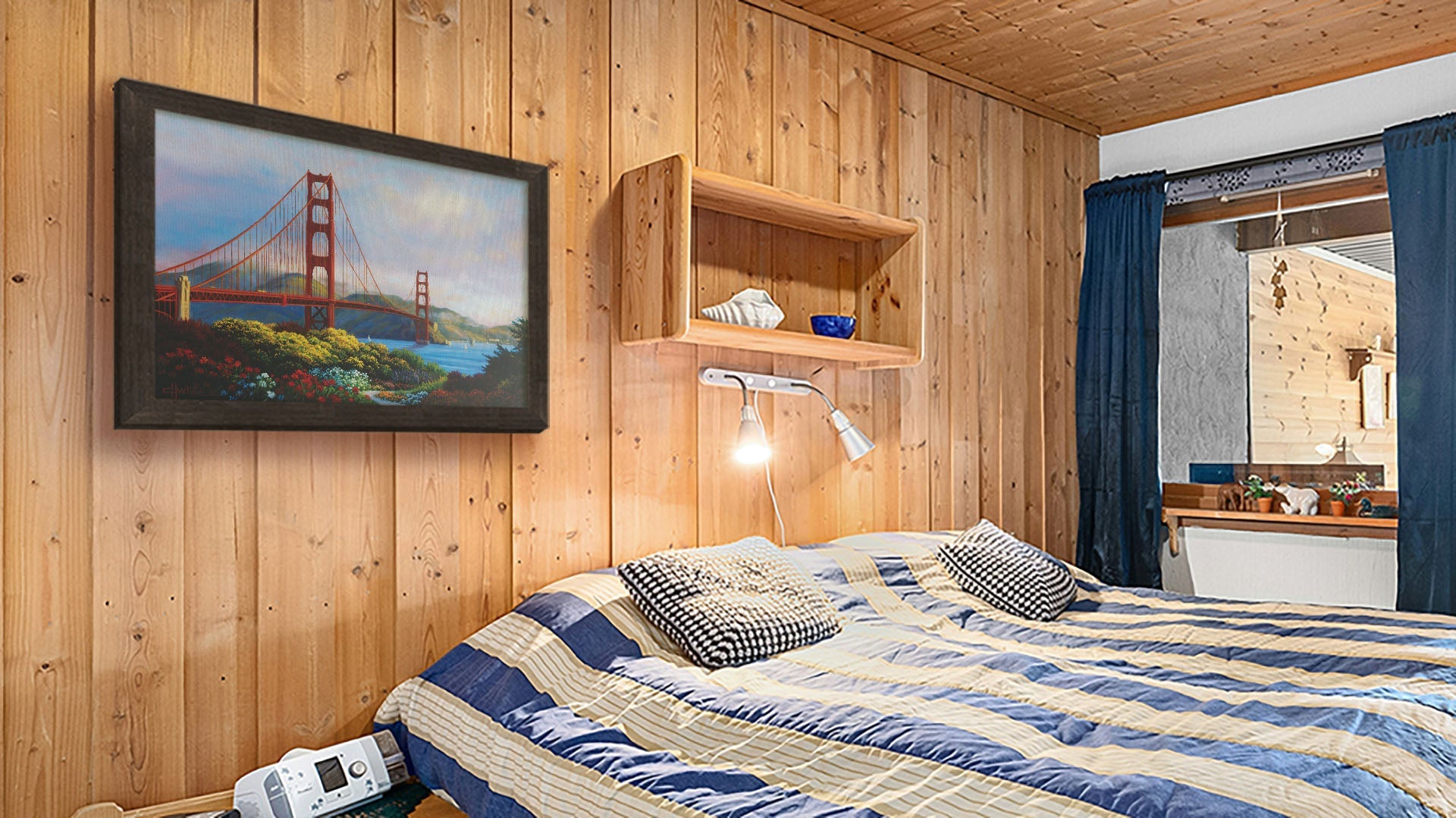 A seaside bedroom with light wood walls and blue and white furniture. A framed painting of the Golden Gate Bridge in San Francisco, California, hangs on the wall.