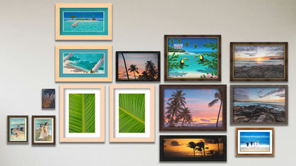 A collage of art in the designer Pastel Palms package, including two matted and framed paintings of kids on the beach; two framed photos of palm trees; a matted and framed photo of kids on the beach; a canvas photo of tropical plants at sunset; two float framed paintings of an island beach and a rowboat at an island; a framed painting of toucans perched on beach plants; two framed photos of Manly Beach, Australia; two framed photos of bird of paradise palms; and a framed photo of a sunset in Maui.