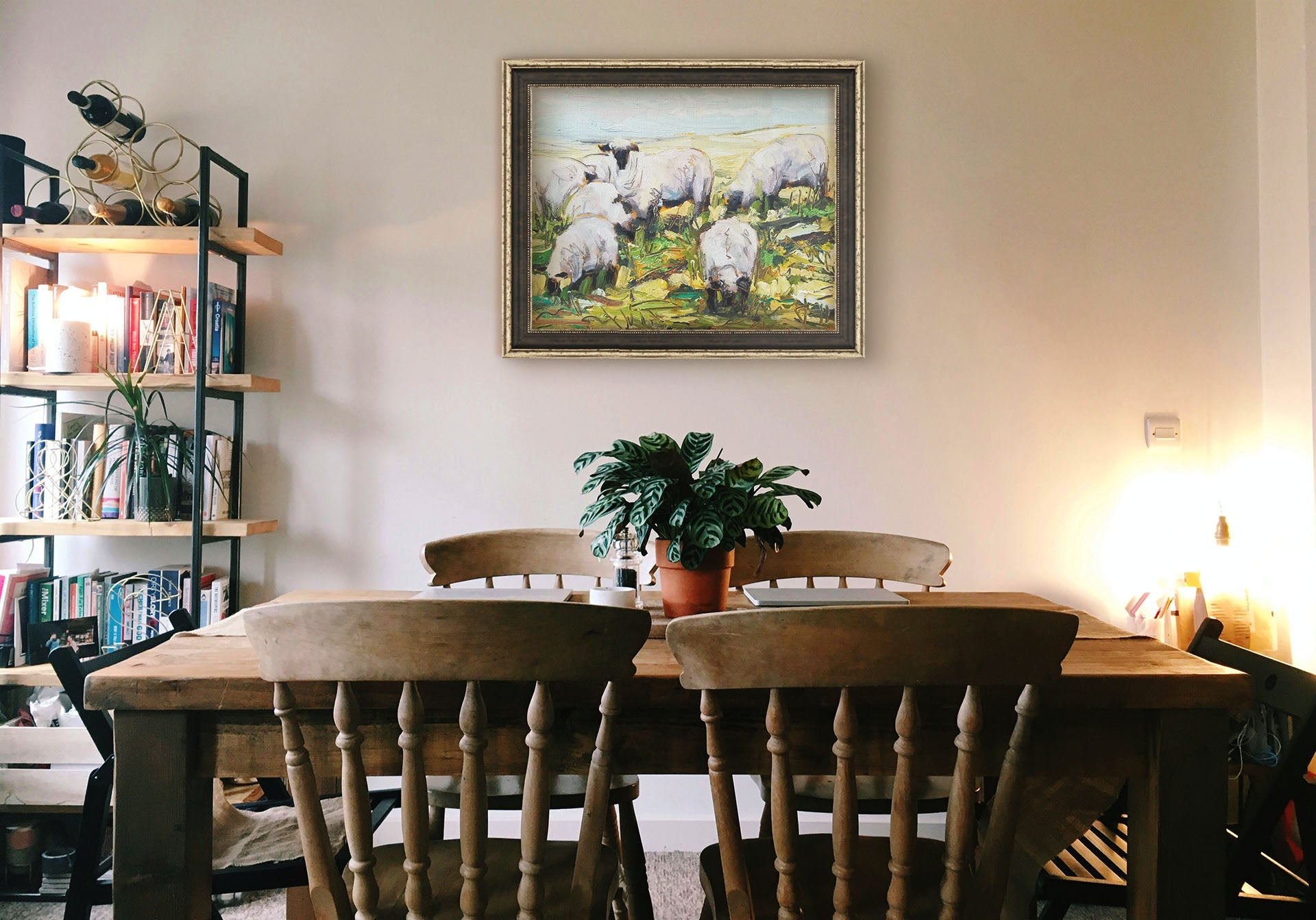 A painting hanging on a dining room wall. A painting of a field of grazing sheep, created, with an expressive impasto style in a palette of soft greens, yellows, and blues. Printed on canvas and framed.