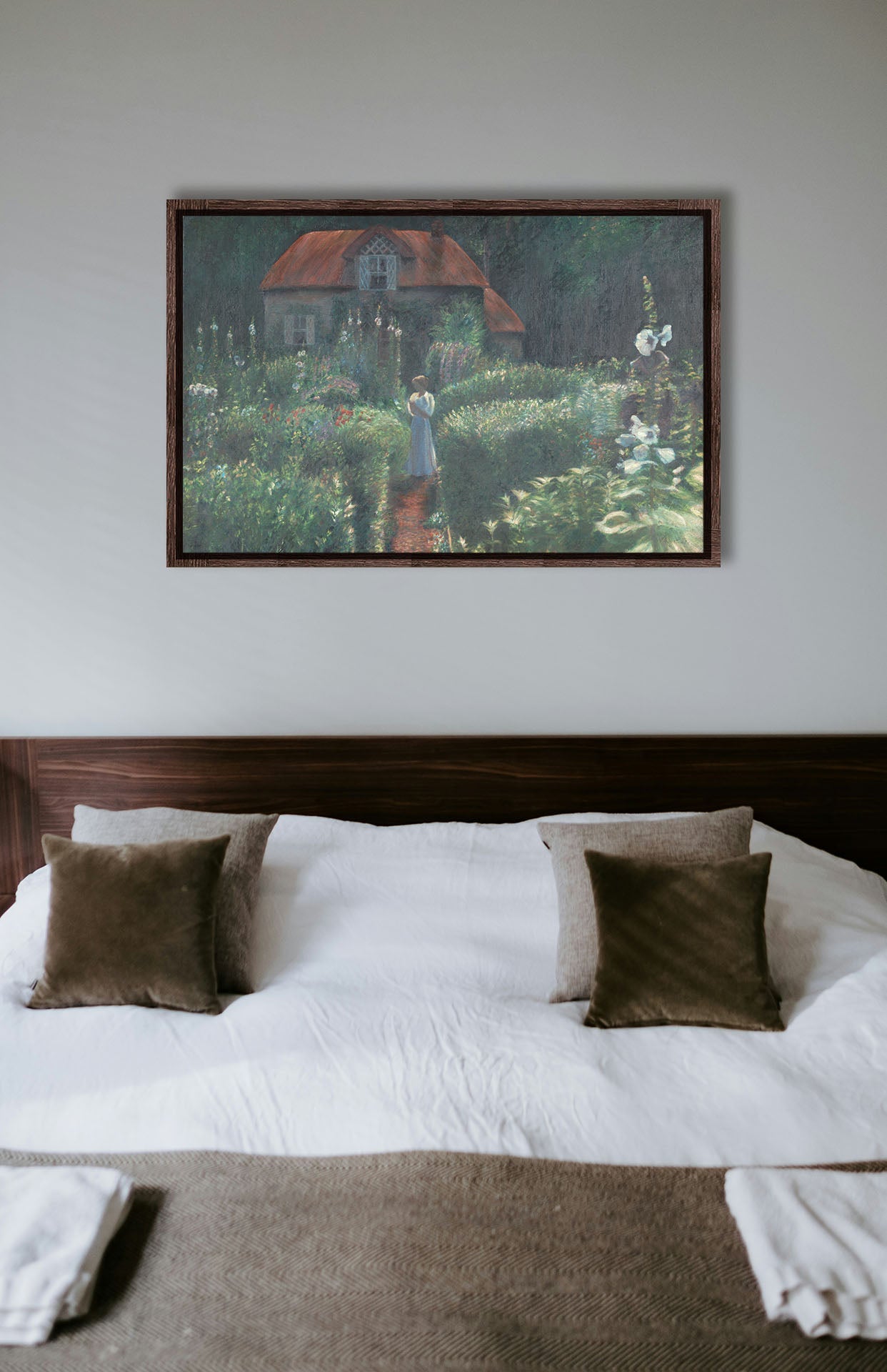 A typical bedroom with light walls and a cozy brown bed. A float framed painting of a cottage surrounded by a garden hangs on the wall.