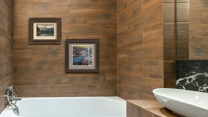 A photograph and a painting hanging on a bathroom wall. The first is a photo overlooking Lake Mamie, one of the Mammoth Lakes at Mammoth Mountain in California. Printed on paper, matted, and framed. The second is a painting of the stone bank of a river, running through a birch forest. Printed on paper, matted, and framed.