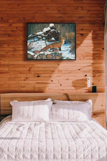 A painting hanging on a bedroom wall. A painting of a red fox in the winter snow. It is standing by some old farming equipment, grasses, and stones. Printed on canvas in a float frame.