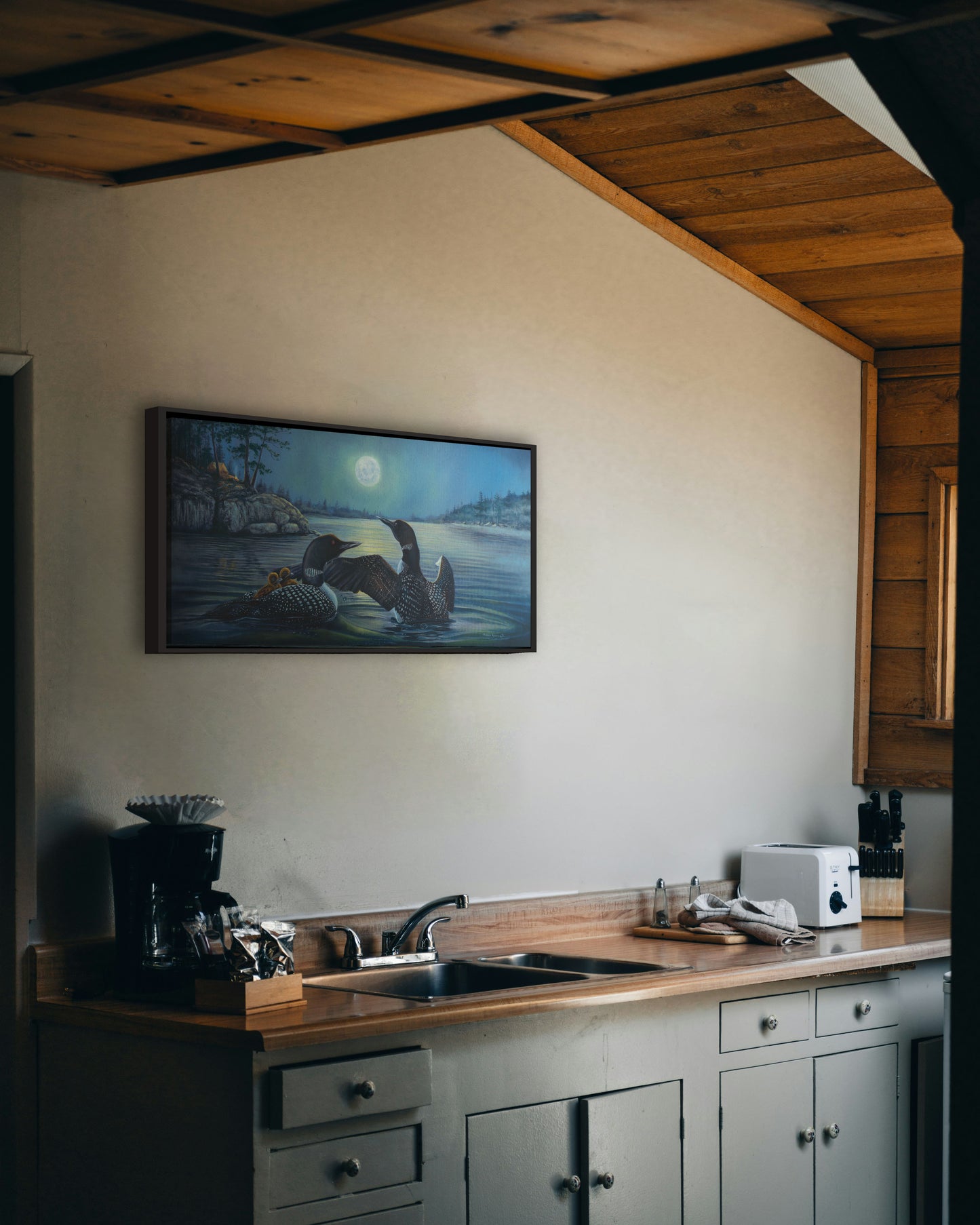 A painting hanging on a kitchen wall. A painting of two grown loons, with two baby loons on one of their backs. They are floating on a lake under the moonlight, with a campground on the land in the distance. Printed on canvas in a float frame.