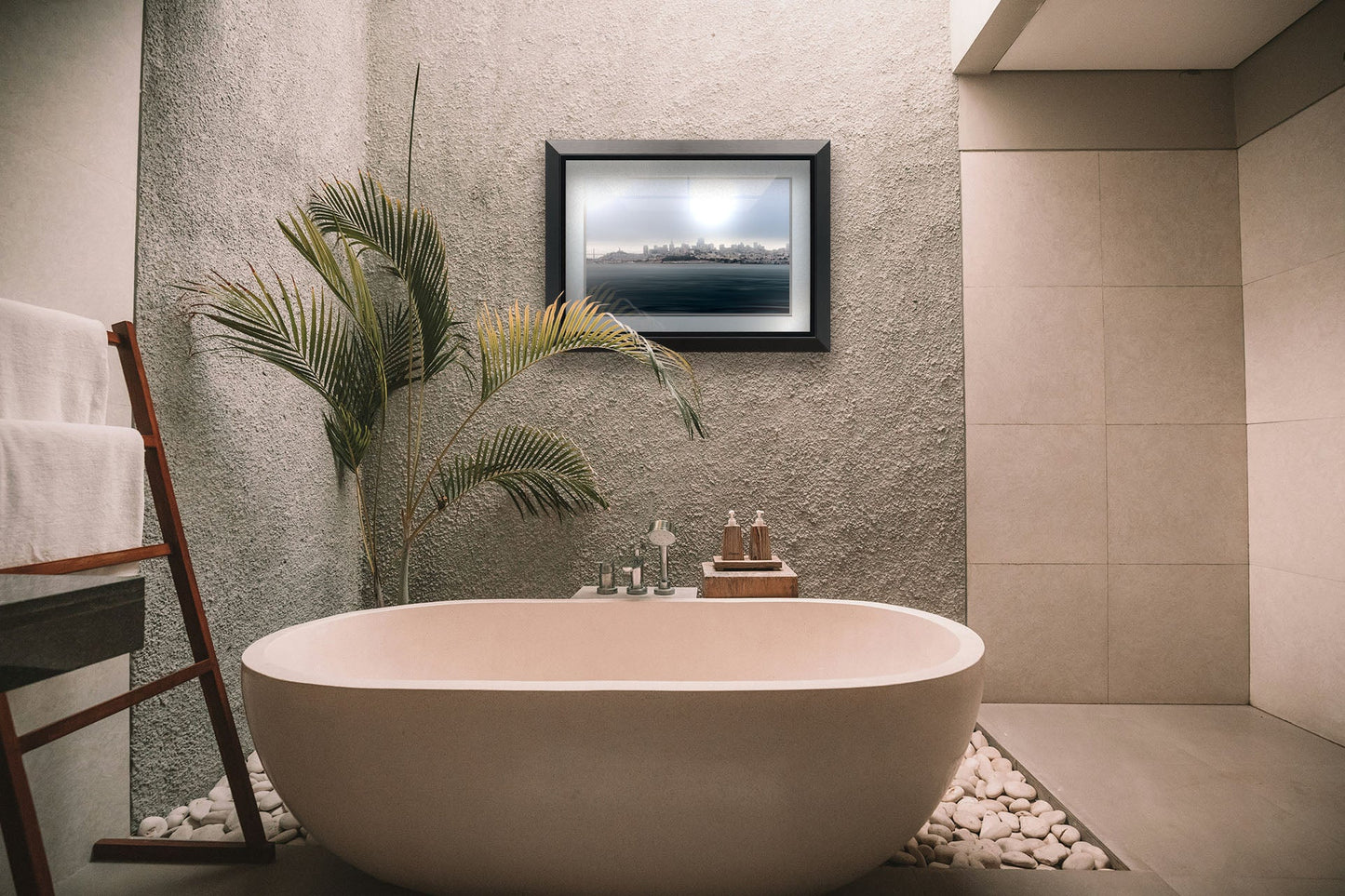 A tropical sea themed bathroom with palm plants and a large tub. A matted and framed photograph overlooking the coast of San Francisco hangs on the wall.