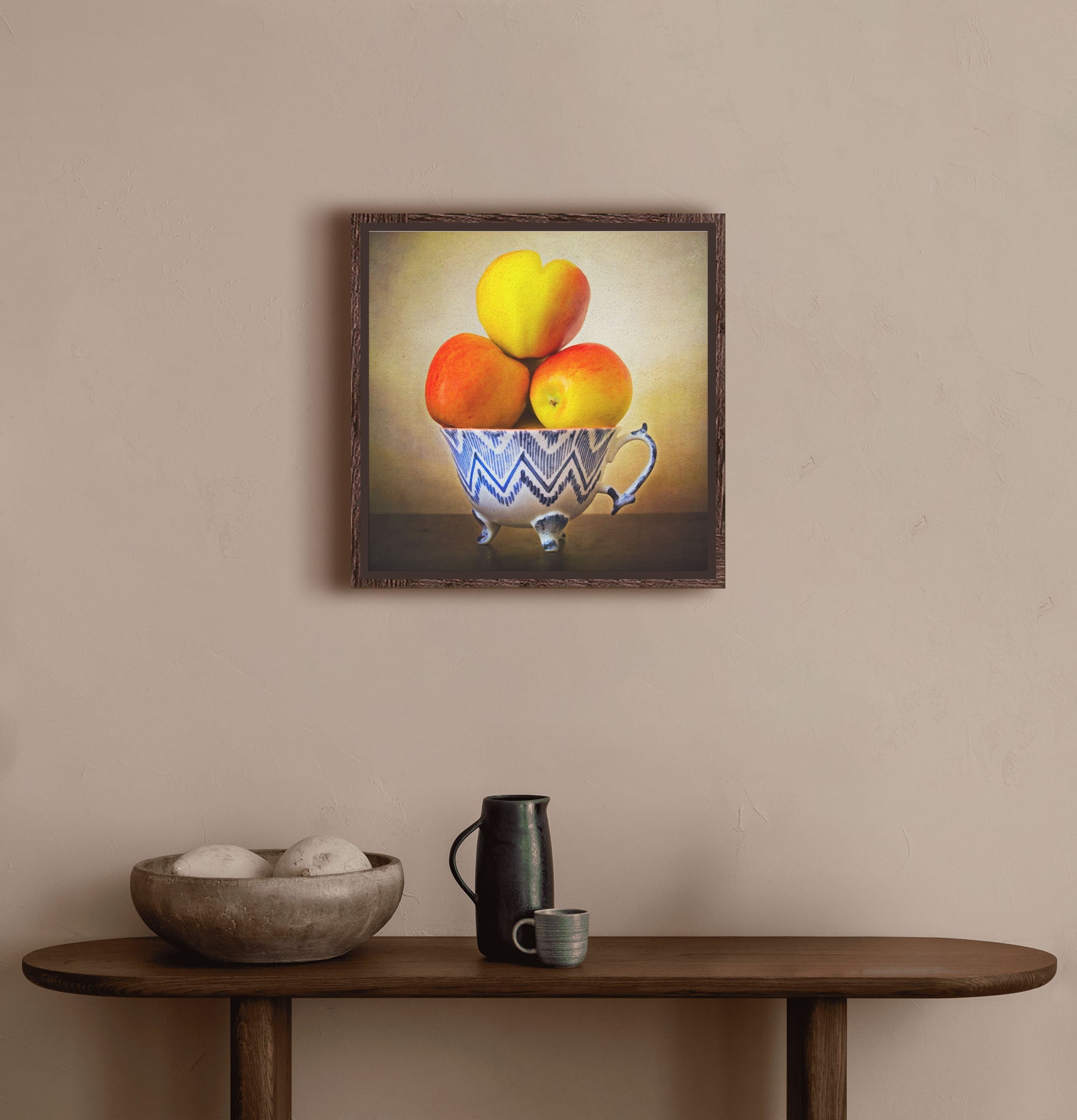 A warm hallway with a small wooden table, with a decorative bowl and ceramics. A float framed photograph of apples stacked in a cup hangs on the wall.