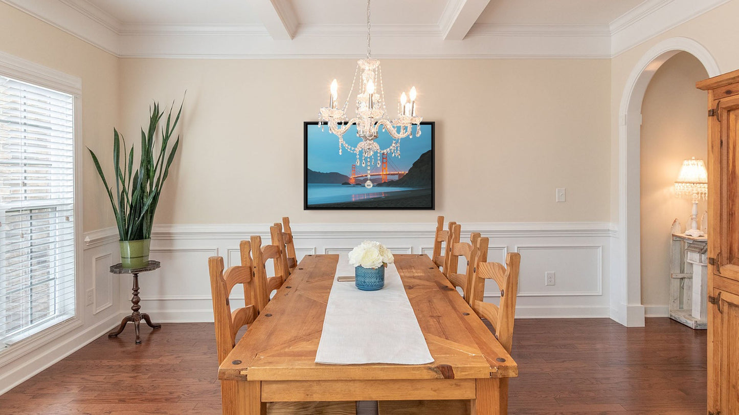 A light dining room with wood furniture, plants, and a clear chandelier. A float framed photograph from Baker Beach, looking out at the Golden Gate Bridge at night.