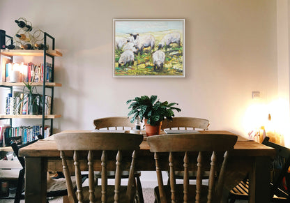 A painting hanging on a dining room wall. A painting of a field of grazing sheep, created, with an expressive impasto style in a palette of soft greens, yellows, and blues. Printed on canvas in a float frame.